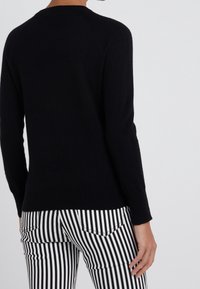 Schwarzer Strickpullover mit Rundhalsausschnitt und langen Ärmeln, kombiniert mit weißen und schwarzen gestreiften Hosen in schmaler Passform.