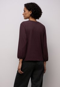 Bordowy sweter z okrągłym dekoltem i rękawami 3/4 w kształcie baloników, wykonany z miękkiej tkaniny, w połączeniu z ciemnymi spodniami.