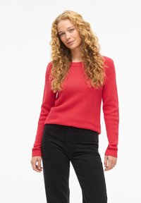 Punainen neulottu paita teksturoidulla kuviolla, pyöreällä pääntiellä ja pitkillä hihoilla, yhdistetty mustiin housuihin. Kangas näyttää pehmeältä ja lämpimältä.