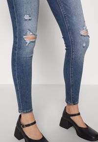 Vaqueros pitillo azules desgastados con roturas en las rodillas, combinados con zapatos de tacón grueso con correa en el tobillo negros y punta redondeada.