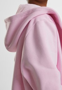 Sudadera con capucha de color rosa claro con una textura suave y cepillada, que presenta una capucha grande con contorno y un ajuste relajado, con detalles de costuras visibles.