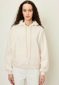 Felpa di pile crema con tasca frontale, cappuccio con lacci e polsini a costine. Indossata con jeans chiari. Tessuto morbido e strutturato.