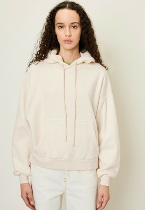 Kremowy polarowy hoodie z kieszenią z przodu, kapturem na sznurki i prążkowanymi mankietami. Noszony z jasnymi dżinsami. Miękka, teksturowana tkanina.