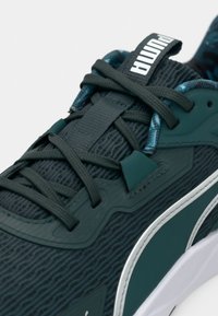 Σκούρο πράσινο αθλητικό παπούτσι Puma με υφασμάτινη υφή, κορδόνια στο ίδιο χρώμα, λευκή σόλα και ορατό λογότυπο Puma στη γλώσσα και την πλευρά.