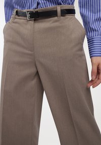 Zbliżenie na osobę ubraną w taupe szerokie spodnie z czarnym paskiem i niebieską koszulę w paski, ręka zrelaksowana przy boku.