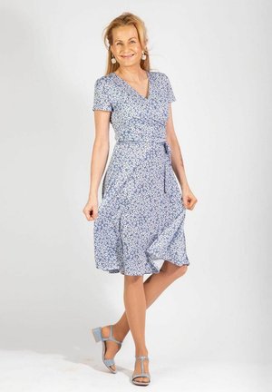 Blaues Blumen-Wickelkleid mit kurzen Ärmeln, einem Bindegürtel in der Taille und einem fließenden Rock. Das Model trägt hellblaue Sandalen. Schlichter Hintergrund.