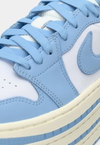 Gros plan d’une basket blanche avec des empiècements en cuir bleu clair, des lacets bleus assortis et une semelle en caoutchouc à rayures bleues et crème.