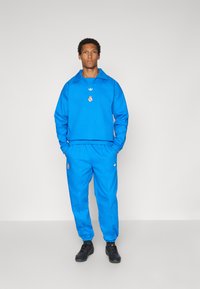 Μπλε φόρμα με μεγάλο γιακά και ελαστική μέση. Διαθέτει λογότυπα στο στήθος και στο παντελόνι. Το υλικό φαίνεται απαλό και με υφή.