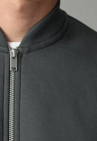 Veste grise à fermeture éclair avec un col côtelé et un tissu texturé, dotée d'une fermeture éclair argentée à l'avant. Design simple et minimaliste.