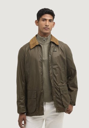 Mann steht und trägt eine olivgrüne Jacke mit braunem Kragen, darunter einen hellgrünen Reißverschluss-Pullover und weiße Hose vor einem schlichten Hintergrund.