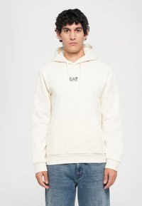 Kremfarget hettegenser i myk stoff med en frontlomme, snor til hetta, og en liten logo på brystet. Brukt sammen med blå jeans.
