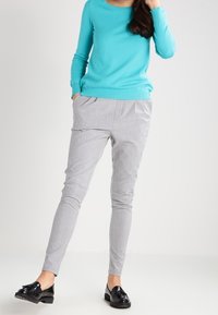 Light gray tapered pants with pleats, paired with a turquoise long-sleeve top. Black loafers complete the outfit. Smooth fabric texture.