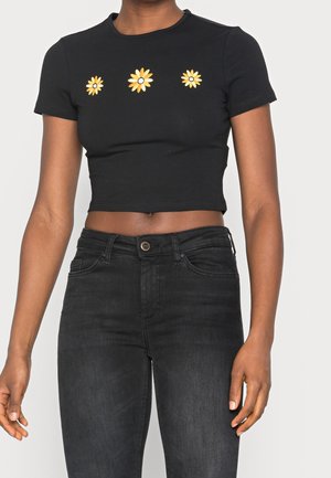 Frau trägt ein schwarzes, kurzes T-Shirt mit drei gelben Gänseblümchen-Motiven auf der Brust und hoch taillierte schwarze Jeans.