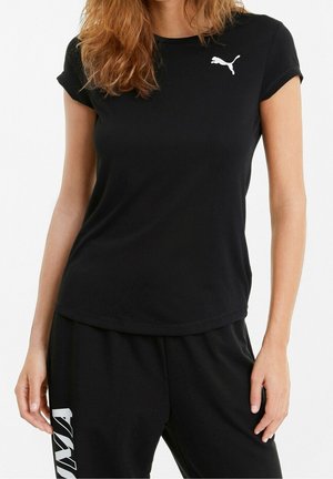 Femme portant un t-shirt noir Puma avec le logo blanc Puma sur la poitrine et un pantalon noir avec un motif blanc sur la cuisse gauche, debout devant un fond blanc.