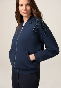 Sudadera azul marino con cierre de cremallera, dos bolsillos frontales y un gran diseño de hoja con lentejuelas en el hombro izquierdo. Textura suave.