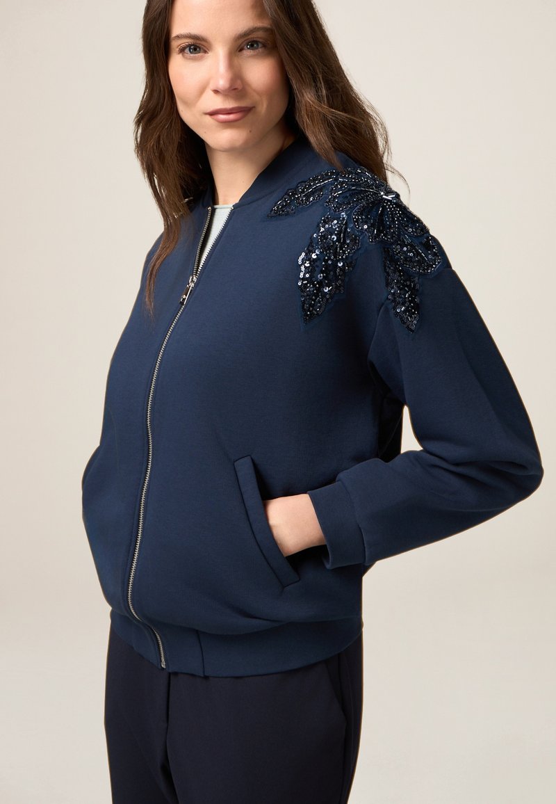 Marineblauwe sweatshirt met rits, twee voorzakken en een groot, geborduurd bladontwerp op de linkerschouder. Zachte textuur.