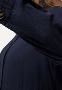 Person som bär en mörkblå långärmad tröja, armen utsträckt framåt, med närbildsfokus på tygtexturen och knappdetaljerna.