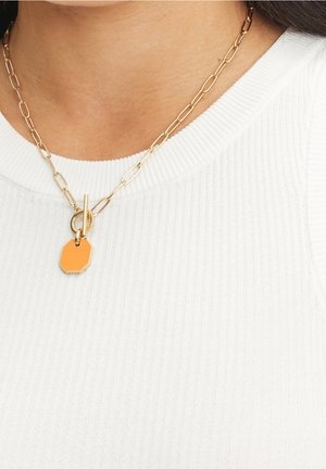 Collier chaîne en or à maillons allongés avec un fermoir à bascule et un pendentif octogonal orange porté sur un haut blanc côtelé.