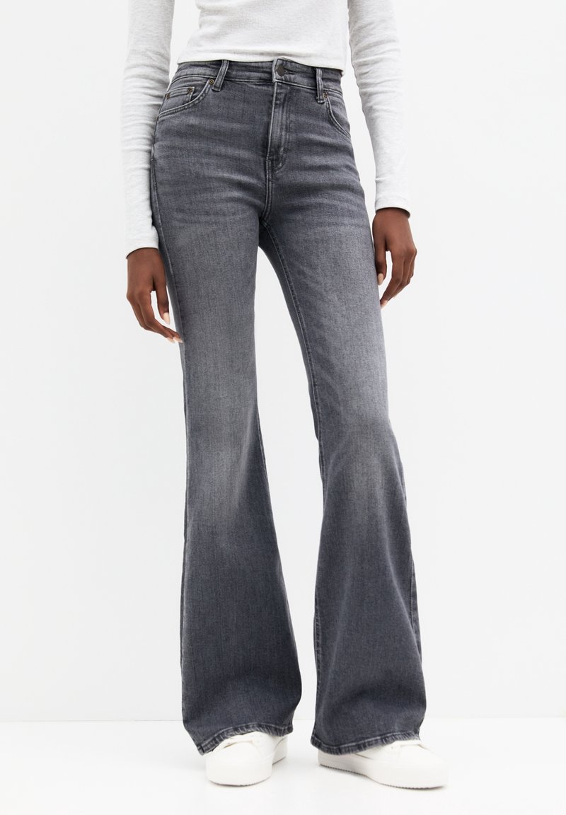 Personne portant un jean évasé gris délavé taille haute, un haut blanc à manches longues et des baskets blanches, debout devant un fond blanc uni.