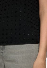 Schwarzes Oberteil mit einem strukturierten Rautenmuster, das kleine Pailletten und florale Akzente aufweist, kombiniert mit grauen Denim-Hosen.