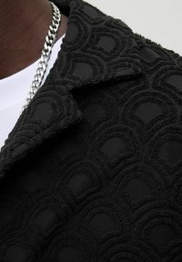 Black textured jacket featuring a raised pattern of interlocking semicircles, with a contrasting white shirt underneath and a silver chain.