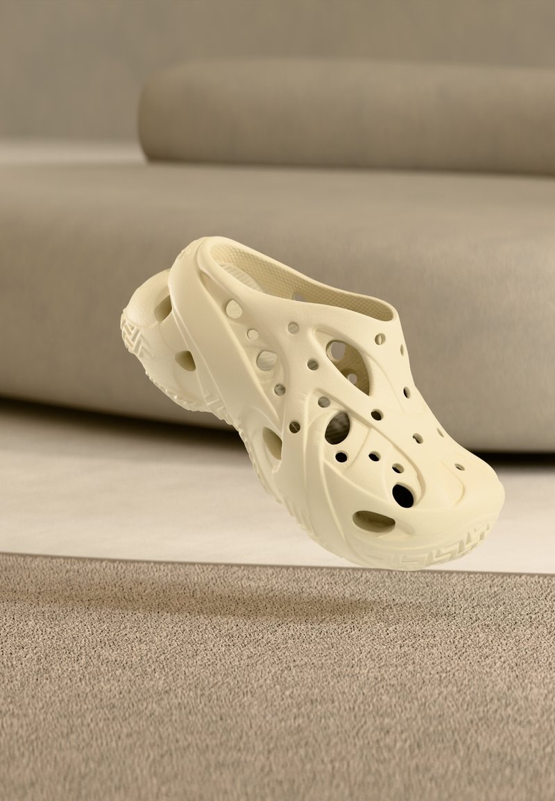 Ζευγάρι κρεμ χρώματος clog χωρίς κορδόνια με πολλές οπές αερισμού, τοποθετημένα σε μπεζ χαλί κοντά σε ανοιχτό καφέ καναπέ.