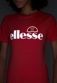 Červené bavlněné tričko s výrazným bílým logem "ellesse" a kruhovým akcentem v tmavě červené nad textem. Hladký povrch, krátké rukávy.