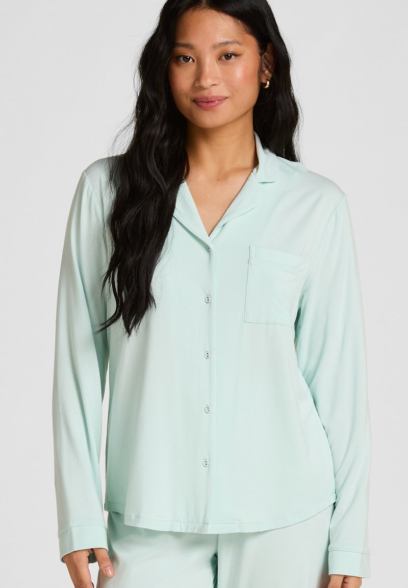 Frau mit langen schwarzen Haaren, die ein hellblaues Pyjamahemd mit Knöpfen und passende Hosen trägt, steht vor einem schlichten Hintergrund.