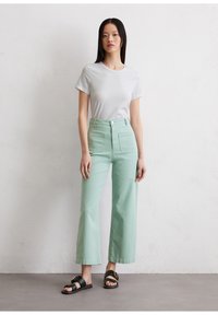 T-shirt blanc à manches courtes associé à un pantalon large taille haute de couleur vert menthe. Sandales noires plates avec un détail en boucle dorée. Tissu lisse et décontracté.