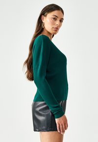 Maglione verde a coste con scollo a V e maniche lunghe, abbinato a pantaloni corti in pelle nera. La texture è liscia con un design aderente.