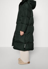 Lang, mørkegrøn pufferjakke med hætte, der har quiltede sektioner, en lynlås foran og sidelommer. Glat, blankt stof.