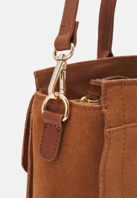 Bruine suède handtas met gouden hardware, voorzien van een afneembare schouderband en een gestructureerd oppervlak. Zakken aan de zijkant zichtbaar met stiksels.