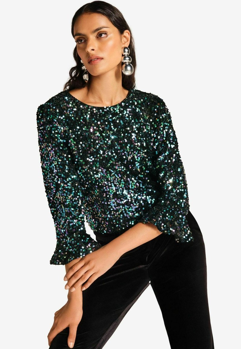 Top in paillettes nero e verde con maniche a tre quarti e polsini arricciati. Outfit abbinato a pantaloni di velluto neri e grandi orecchini argento.