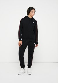 Conjunto de sudadera con capucha y pantalones negros hechos de tela suave, con rayas rojas a lo largo de las mangas y las piernas, y detalles de logotipo en el pecho y el muslo.