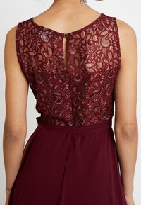 Ärmlös klänning i vinröd med blommig spets på ryggen och en liten knappknäppning i mitten på toppen, bärs av en person med tatueringar på armarna.