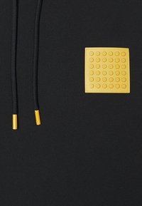 Primer plano de una tela negra con cordones negros que tienen puntas amarillas y un parche cuadrado amarillo con puntos en relieve en la zona del pecho.