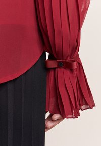 Rød gennemsigtig bluse med plisserede ærmer, der har en satinbånd detalje og knaplukning. Matchende med et ensfarvet sort stof forneden.