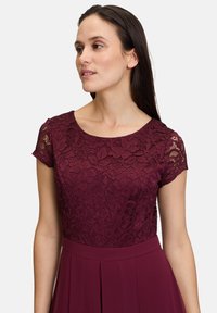 Burgunderfarbenes Kleid mit einem Spitzenoberteil mit floralen Mustern und kurzen Ärmeln, das in einen plissierten Rock übergeht, glatte Stoffstruktur.