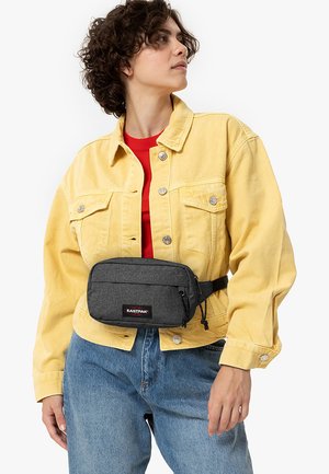 Riñonera gris de Eastpak llevada sobre una chaqueta de mezclilla amarilla. La riñonera cuenta con un cierre de cremallera y una etiqueta de marca en la parte delantera.