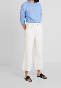 Camicia button-up azzurro chiaro con taschino sul petto, abbinata a jeans bianchi a gamba larga con orli sfrangiati e sandali neri a scivolo.