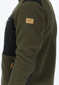Olivgrüne Fleecejacke mit schwarzen Einsätzen, strukturierter Oberfläche, Reißverschlusstaschen und einem Lederlogopatch am Ärmel.