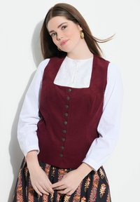 Gilet borgogna con colletto in pizzo bianco, dotato di bottoni neri frontali. Indossato sopra una gonna a fantasia con accenti floreali.