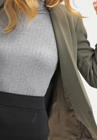 Olive green blazer with textured fabric, worn over a light gray ribbed top and a black high-waisted skirt featuring decorative stitching.