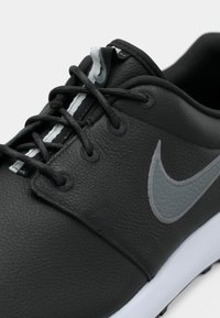 Μαύρο αθλητικό παπούτσι Nike με μαύρα κορδόνια, γκρι λογότυπο Swoosh και λευκή σόλα, εμφανιζόμενο σε κοντινή γωνιακή λήψη σε λευκό φόντο.