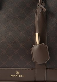 Barna kézitáska texturált külsővel, arany színű kiegészítőkkel és "ANNA FIELD" márkajelzéssel ellátott címkével. A dizájn finom mintázattal rendelkezik az egész felületen.