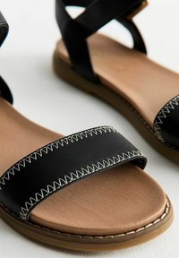 Sandalias de piel negra con una tira ancha que presenta costuras blancas, una plantilla color tostado y una suela marrón flexible. Tiras cruzadas en el tobillo.