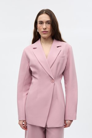 Žena s dlhými hnedými vlasmi, ktorá má na sebe na mieru ušitý svetloružový blazer s jedným gombíkom, stojaca pred obyčajným bielym pozadím.
