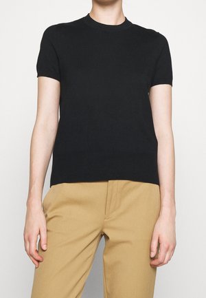 Personne portant un haut en tricot noir à manches courtes associé à un pantalon beige taille haute, debout devant un fond uni.