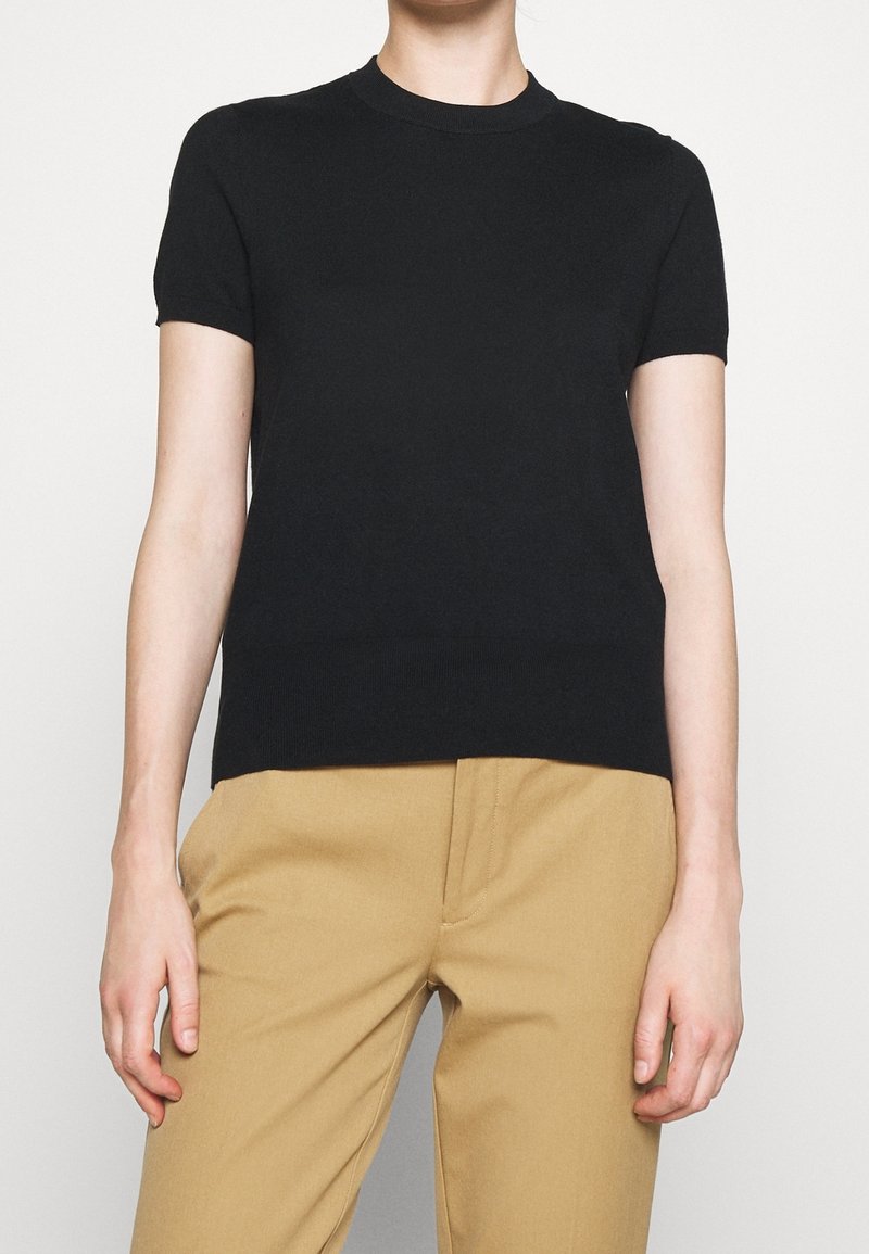 Personne portant un haut en tricot noir à manches courtes associé à un pantalon beige taille haute, debout devant un fond uni.