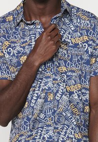 Homme à la peau foncée portant une chemise bleue à manches courtes avec du texte jaune et blanc et des motifs floraux, ajustant le col avec sa main droite.